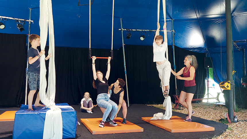 Des enfants essayent des acrobaties avec des tissus alors qu'au centre de l'image une accompagnatrice aide une jeune fille à faire une figure au trapèze