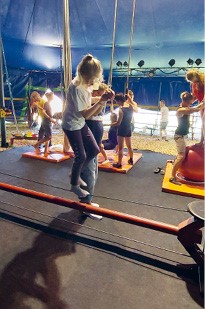 Sous le chapiteau du cirque, une jeune fille est en équilibre sur un fil, soutenue par un accompagnateur 